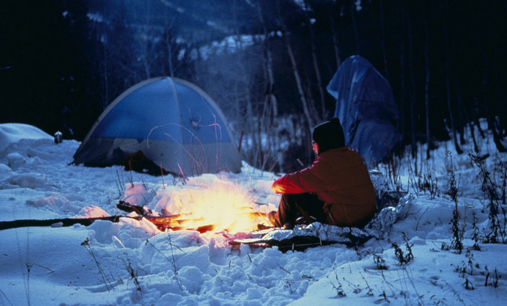 A person sat in the snow with a campfire infront of them, and a tent in the background