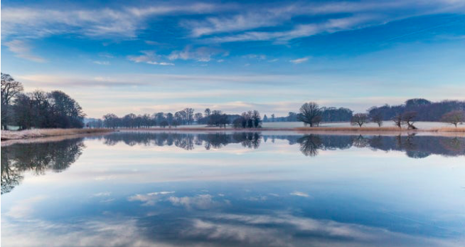 A beautiful blue lake with trees reflected in it with blue sky.