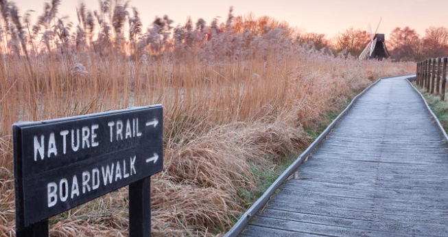 A field with tall brown grass with a wooden pathway on one side. There is a sign which reads "Nature Trail and Boardwalk"