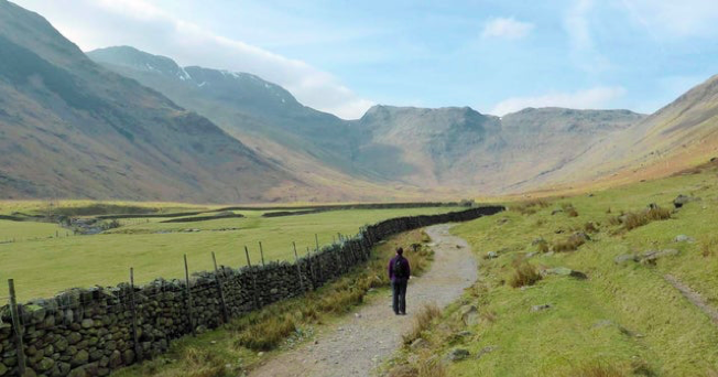 A field of green grass with a man walking down a path in the middle, there is a cobbled wall to one side and a group of green hills in the background against a blue sky. 