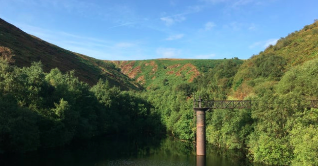 A blue sky and green hills with trees fill the image, with a small, brown, metal bridge on one side.