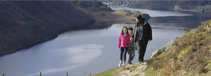 A woman and two children walking along a path on a mountain, and there is a lake in the background