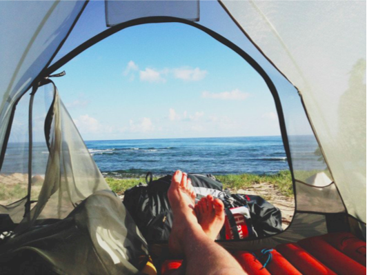 This image has been taken by someone lying down in a tent, showing the view outside the tent door, looking out at a line of grass and then the blue sea.