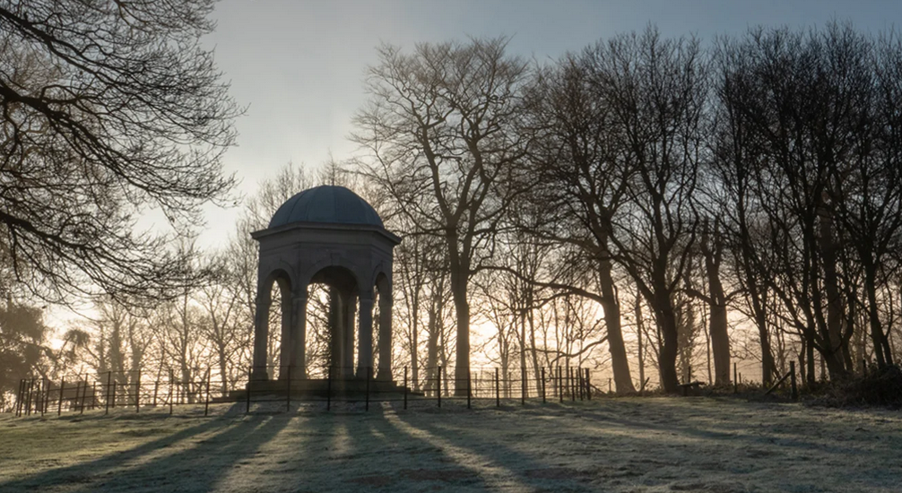 A field with frost covered grass, surrounded by tall, leafless trees with the sum shining through them. In the middle of the image is a small dome shaped buidling. 