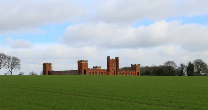 A large, green field with blue sky. There is a very large orange brick castle-type bulding in the field.