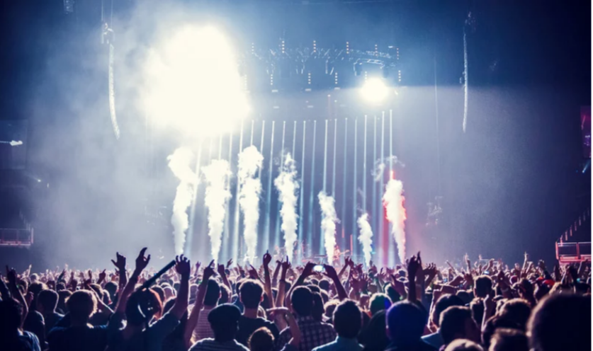 An image of a festival. There is a large group of people infront of a stage who are dancing with their arms in the air. There is smoke coming from the stage.