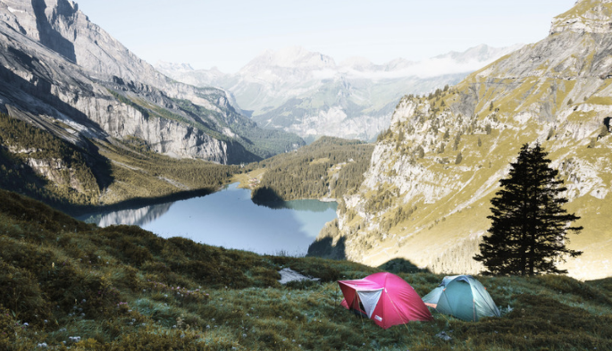 A red and blue tent, pitched by a beautiful blue lake which is surrounded by rocky mountains and trees. 