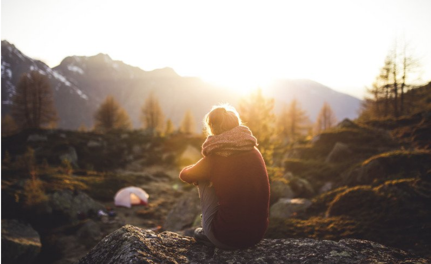 A woman facing away, wearing a scarf, sat on a rock. She is overlooking trees with her tent set up in the background. The sunshine is beaming down on her.