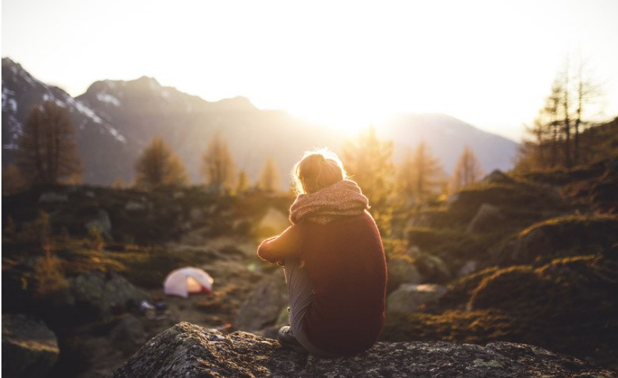 A woman facing away, wearing a scarf, sat on a rock. She is overlooking trees with her tent set up in the background. The sunshine is beaming down on her. 