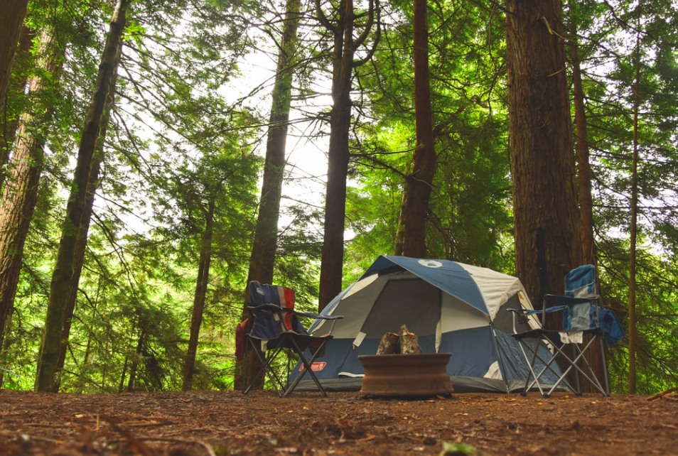 A blue tent in the woods with two camping chairs infront of it.