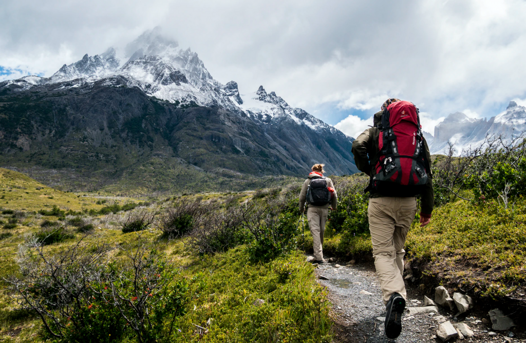 Two people with backpacks walking along a grassy path towards mountains