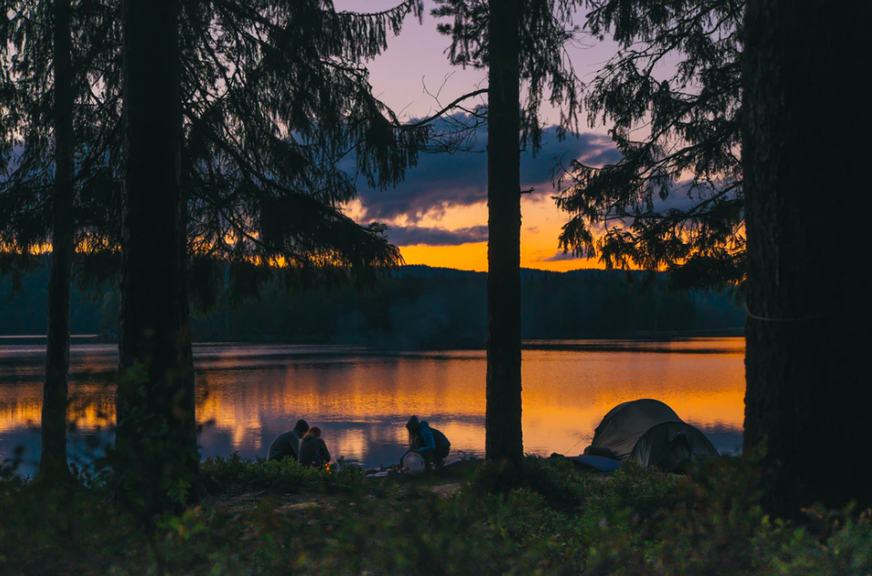 Three people sitting by a lake with their tent next to them during sunset